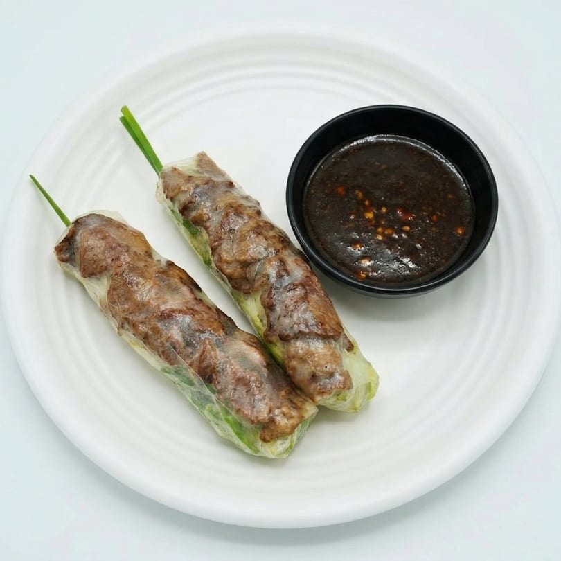 Two fresh spring rolls with beef and green herbs, served with a dark dipping sauce on a white plate.